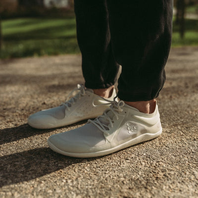 A photo of Vivobarefoot Primus lite sneakers made from recycled plastic and a rubber sole. The athletic sneakers are a white color with white soles and the vivobarefoot logo on the side. Shoes are shown facing left on a person wearing black joggers standing on a paved road with grass in the background. #color_bright-white