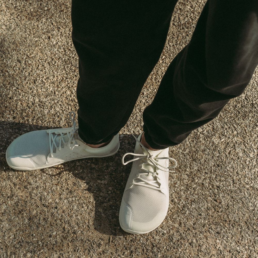 A photo of Vivobarefoot Primus lite sneakers made from recycled plastic and a rubber sole. The athletic sneakers are a white color with white soles and the vivobarefoot logo on the side. Shoes are shown from above on a person wearing black joggers standing on a paved road with grass in the background. #color_bright-white