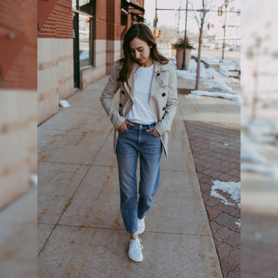 A photo of Vivobarefoot Primus lite sneakers made from recycled plastic and a rubber sole. The athletic sneakers are a white color with white soles and the vivobarefoot logo on the side. Shoes are shown from the front on a tan woman with long brown hair wearing a white shirt tucked into loose-fitting jeans and a tan trench coat. She is walking on a sidewalk with her hands in her pockets and an urban setting in the background. #color_bright-white