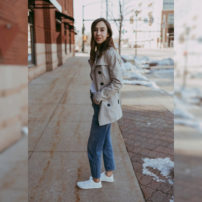 A photo of Vivobarefoot Primus lite sneakers made from recycled plastic and a rubber sole. The athletic sneakers are a white color with white soles and the vivobarefoot logo on the side. Shoes are shown facing left on a tan woman with long brown hair wearing a white shirt tucked into loose-fitting jeans and a tan trench coat. She is standing on a sidewalk with her hands in her pockets and an urban setting in the background. #color_bright-white