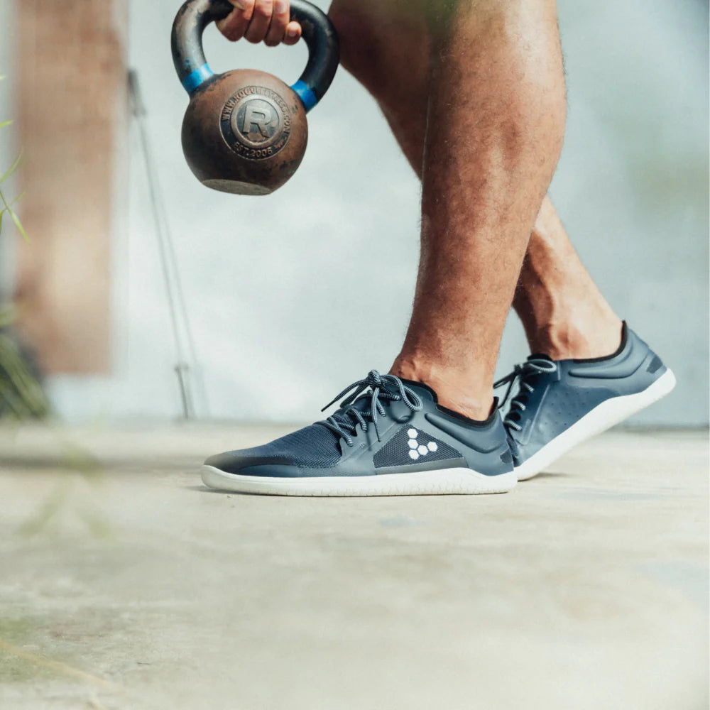 A photo of recycled plastic Navy Vivobarefoot Primus lite sneakers with white rubber soles and a white vivobarefoot logo on the side. Recycled mesh fabric surrounds the shoe with a thicker recycled guard around the sole, laces, and ankle opening. Both shoes are shown facing left on a man holding a kettlebell doing a left-legged hinge on a cement ground. #color_navy