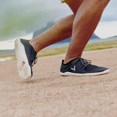 A photo of recycled plastic Navy Vivobarefoot Primus lite sneakers with white rubber soles and a white vivobarefoot logo on the side. Recycled mesh fabric surrounds the shoe with a thicker recycled guard around the sole, laces, and ankle opening. Both shoes are shown facing right on a man doing a speed skater movement on his right leg exposing the left shoe sole on a sand ground with a mountain in the background. #color_navy