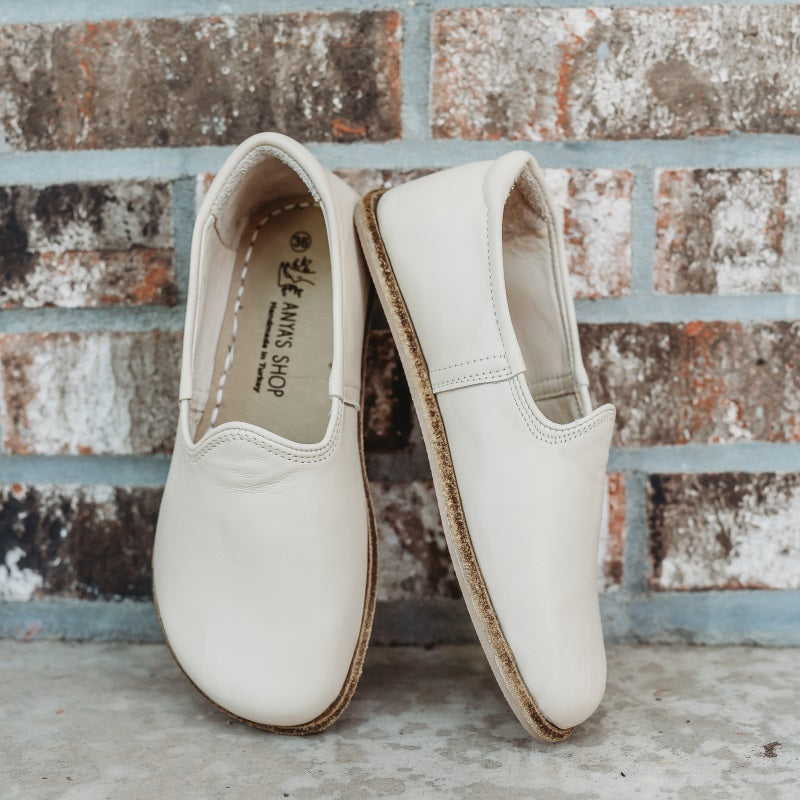 A photo of Yasemin Leather loafers Designed by Anya with a leather upper and tan rubber soles. The loafers are a smooth cream/beige color and have a small curve up on the top of the foot for design. Both loafers are shown leaning against a brick wall with the right shoe facing to the right side both resting on cement. #color_beige