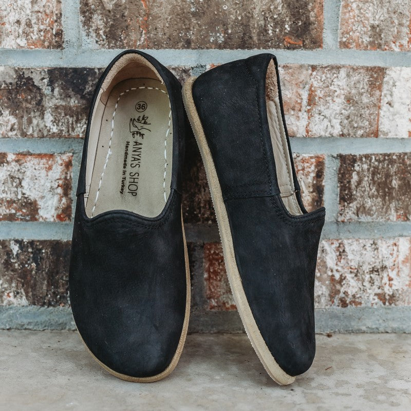 A photo of Yasemin Leather loafers Designed by Anya with a leather upper and tan rubber soles. The loafers are a black nubuck color and have a small curve up on the top of the foot for design. Both loafers are shown leaning against a brick wall with the right shoe facing to the right side both resting on cement. #color_black-nubuck