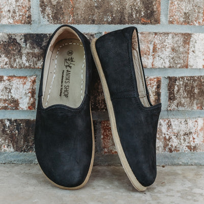 A photo of Yasemin Leather loafers Designed by Anya with a leather upper and tan rubber soles. The loafers are a black nubuck color and have a small curve up on the top of the foot for design. Both loafers are shown leaning against a brick wall with the right shoe facing to the right side both resting on cement. #color_black-nubuck