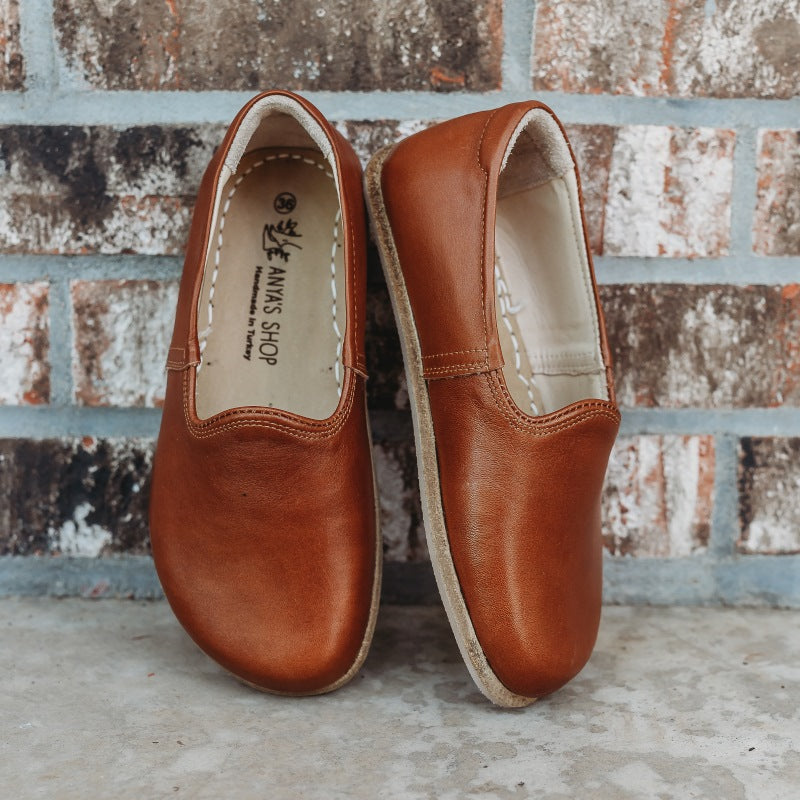 A photo of Yasemin Leather loafers Designed by Anya with a leather upper and tan rubber soles. The loafers are a smooth brown color and have a small curve up on the top of the foot for design. Both loafers are shown leaning against a brick wall with the right shoe facing to the right side both resting on cement. #color_brown
