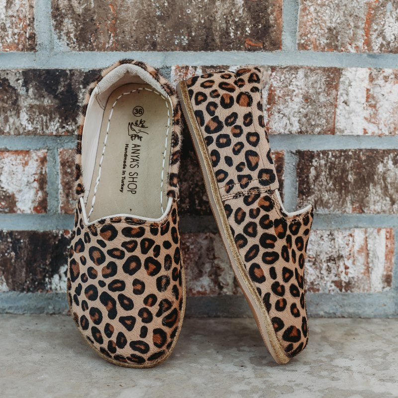 A photo of Yasemin Leather loafers Designed by Anya with a leather upper and tan rubber soles. The loafers are a leopard print and have a small curve up on the top of the foot for design. Both loafers are shown leaning against a brick wall with the right shoe facing to the right side both resting on cement. #color_leopard-print-calf-hair