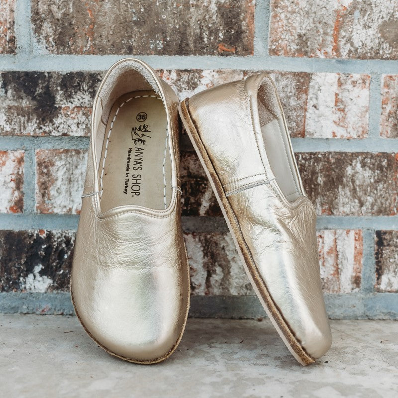 A photo of Yasemin Leather loafers Designed by Anya with a leather upper and tan rubber soles. The loafers are a metallic gold color and have a small curve up on the top of the foot for design. Both loafers are shown leaning against a brick wall with the right shoe facing to the right side both resting on cement. #color_gold