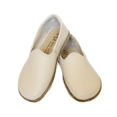 A photo of Yasemin Leather loafers Designed by Anya with a leather upper and tan rubber soles. The loafers are a smooth beige leather upper and have a small curve up on the top of the foot for design. Both loafers are shown from the front with the heel of the right shoe resting on the ankle opening of the left shoe against a white background. #color_beige