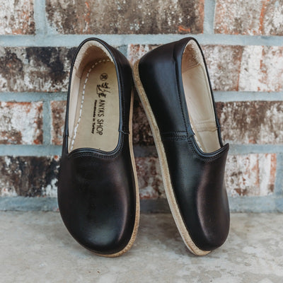 A photo of Yasemin Leather loafers Designed by Anya with a leather upper and tan rubber soles. The loafers are a smooth black color and have a small curve up on the top of the foot for design. Both loafers are shown leaning against a brick wall with the right shoe facing to the right side both resting on cement. #color_black-smooth-leather