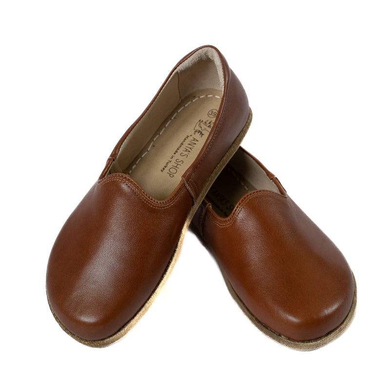 A photo of Brown Yasemin Leather loafers Designed by Anya with a leather upper and tan rubber soles. The loafers have a small curve up on the top of the foot for design. Shoe is shown with the left heel stacked on top of the right shoe opening against a white background. #color_brown