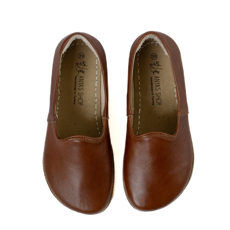 A photo of Brown Yasemin Leather loafers Designed by Anya with a leather upper and tan rubber soles. The loafers have a small curve up on the top of the foot for design. Shoes are shown from the top down against a white background. #color_brown