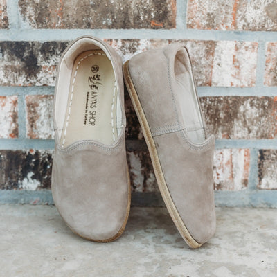 A photo of Yasemin Leather loafers Designed by Anya with a leather upper and tan rubber soles. The loafers are a taupe nubuck color and have a small curve up on the top of the foot for design. Both loafers are shown leaning against a brick wall with the right shoe facing to the right side both resting on cement. #color_taupe-nubuck