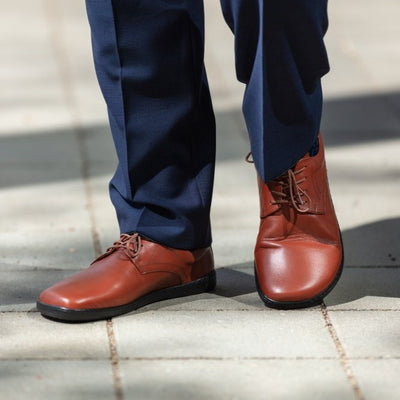 Cognac Zaqq Peaq leather dress shoes with coganc laces and black rubber soles. Both shoes are shown here. The right shoe is facing diagonally left and the right is facing front propped up on a person wearing navy dress slacks standing on a square paver path. #color_cognac