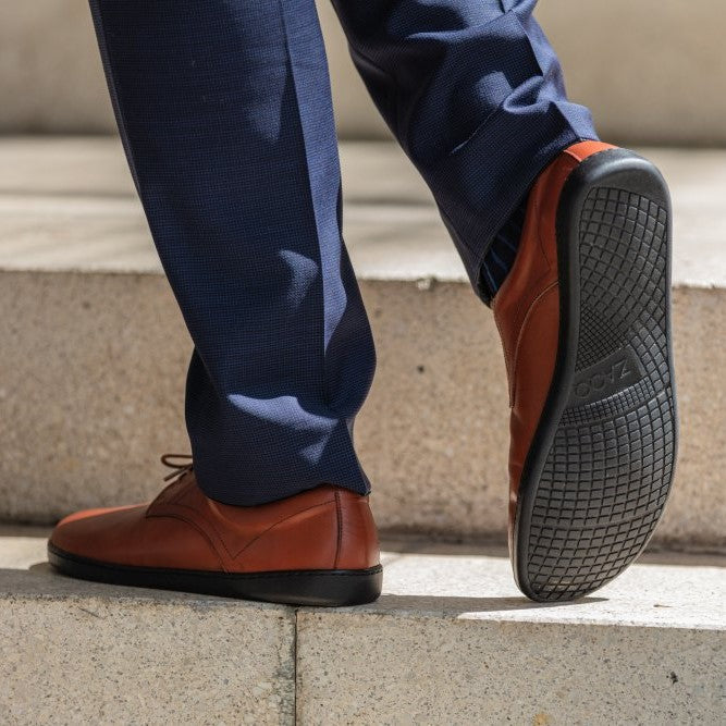 Cognac Zaqq Peaq leather dress shoes with coganc laces and black rubber soles. Both shoes are shown here. The right left shoe is facing to the back left diagonal while the right shoe sole is propped up on a person wearing navy slacks standing on stone stairs. #color_cognac