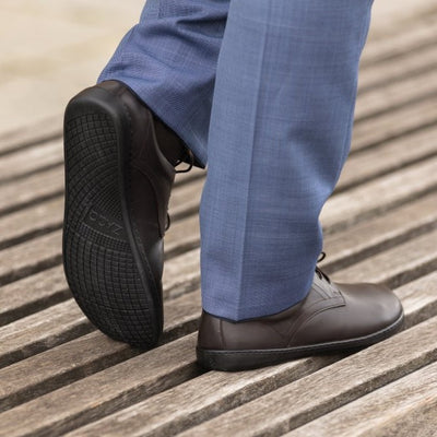 Dark Brown Zaqq Peaq leather dress shoes with dark brown laces and black rubber soles. Both shoes are shown from the diagonal right back with the left foot propped up showing the sole on a person wearing blue dress slacks standing on slatted weathered wood. #color_dark-brown