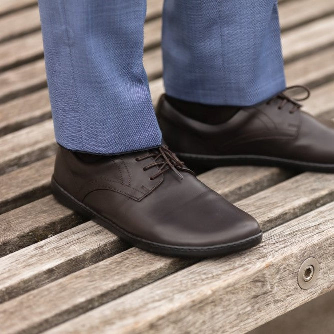 Dark Brown Zaqq Peaq leather dress shoes with dark brown laces and black rubber soles. Both shoes are shown facing diagonally right on a person wearing blue dress slacks standing on slatted weathered wood. #color_dark-brown