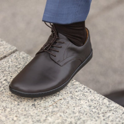 Dark Brown Zaqq Peaq leather dress shoes with dark brown laces and black rubber soles. Left shoe is shown facing diagonally left propped up on a stone wall on a person wearing dark brown socks and blue dress slacks. #color_dark-brown