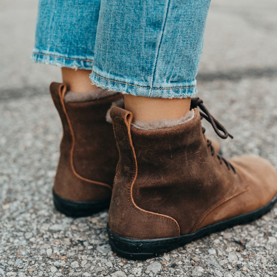 A photo of Zaqq Quintic boots made nappa leather, wool, and rubber soles. The boots are brown in color with dark brown laces and a wool lining inside. Both boots are shown here from the back facing diagonally to the right on a woman's feet wearing cropped blue jeans standing on a paved road with gass in the background. #color_brown