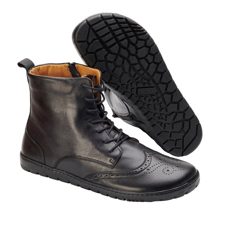 Black Zaqq Quintic Brogue leather, unlined boots with side zippers. Shoes go over the ankles, a pull tab at the top back, have tan lining, and have a brogue design on the toebox. Right shoe is shown facing diagonally right with the left shoe sole propped up on it against a white background. #color_black