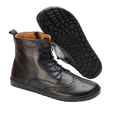 Black Zaqq Quintic Brogue leather, unlined boots with side zippers. Shoes go over the ankles, a pull tab at the top back, have tan lining, and have a brogue design on the toebox. Right shoe is shown facing diagonally right with the left shoe sole propped up on it against a white background. #color_black