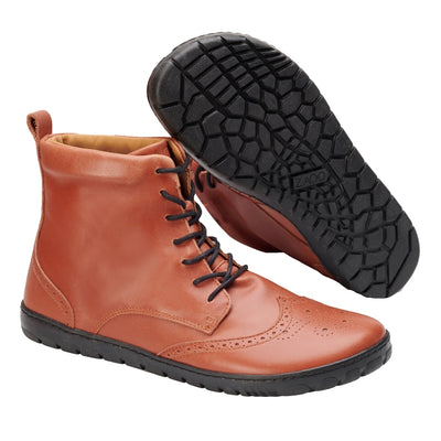 Cognac Zaqq Quintic Brogue leather, unlined boots. Shoes go over the ankles, have black soles and laces, a pull tab at the top back, and have a brogue design on the toebox. Right shoe is shown facing diagonally right with the left shoe sole propped up on it against a white background. #color_cognac