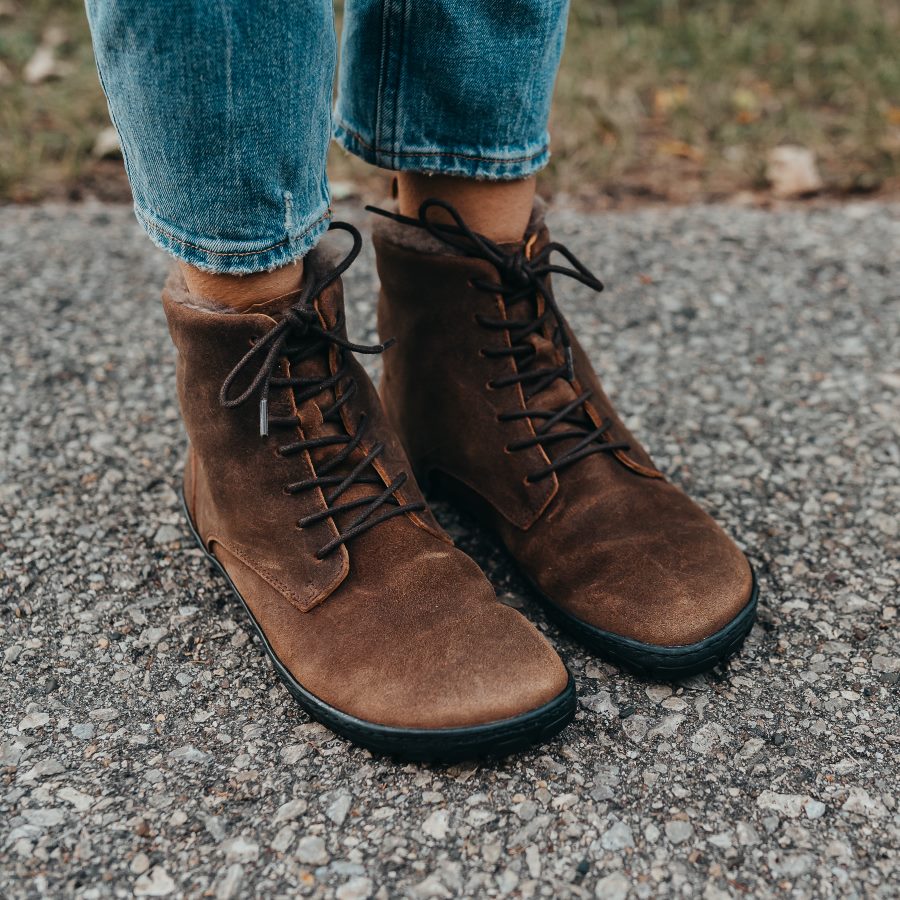 A photo of Zaqq Quintic boots made nappa leather, wool, and rubber soles. The boots are brown in color with dark brown laces and a wool lining inside. Both boots are shown here facing diagonally to the right on a woman's feet wearing cropped blue jeans standing on a paved road with gass in the background. #color_brown
