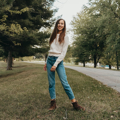 A photo of Zaqq Quintic boots made nappa leather, wool, and rubber soles. The boots are brown in color with dark brown laces and a wool lining inside. Both boots are shown here on a woman's feet wearing cropped blue jeans and a white sweater standing on grass with a paved road and pine trees in the background. #color_brown