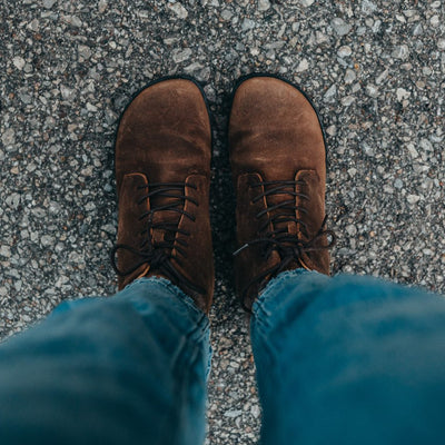 A photo of Zaqq Quintic boots made nappa leather, wool, and rubber soles. The boots are brown in color with dark brown laces and a wool lining inside. Both boots are shown here from above on a woman's feet wearing cropped blue jeans standing on a paved road. #color_brown