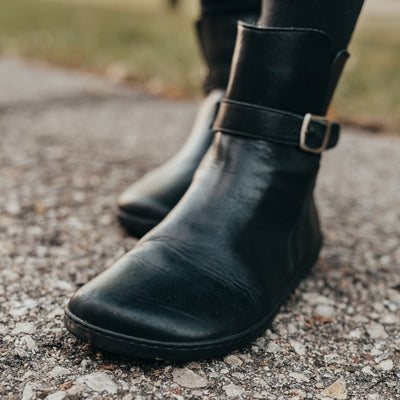 A photo of Zaqq Riquet boots made from Nappa leather and rubber soles. The boots are black in color and a pull up style with a decorative buckle around the ankle. Both boots are shown on a womans feet from the front left wearing black leggings on a paved road with grass in the background. #color_black