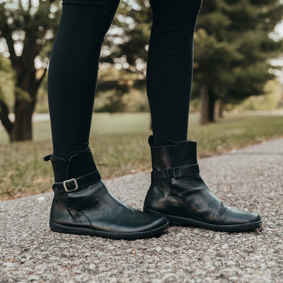 A photo of Zaqq Riquet boots made from Nappa leather and rubber soles. The boots are black in color and a pull up style with a decorative buckle around the ankle. Both boots are shown on a womans feet which are staggered facing right wearing black leggings on a paved road with grass and trees in the background. #color_black