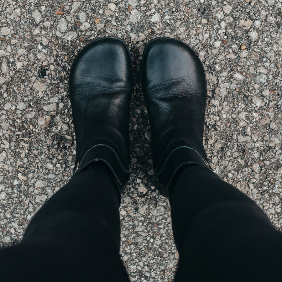 A photo of Zaqq Riquet boots made from Nappa leather and rubber soles. The boots are black in color and a pull up style with a decorative buckle around the ankle. Both boots are shown from the top facing down on a womans feet wearing black leggings standing on a paved road. #color_black