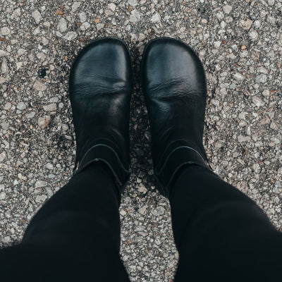A photo of Zaqq Riquet boots made from Nappa leather and rubber soles. The boots are black in color and a pull up style with a decorative buckle around the ankle. Both boots are shown from the top facing down on a womans feet wearing black leggings standing on a paved road. #color_black