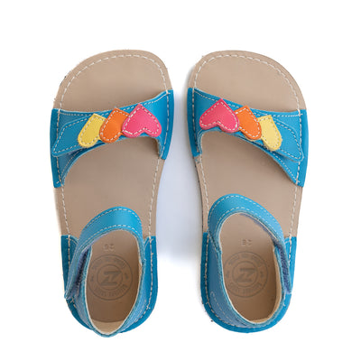 A photo of the Zeazoo Ariel kids leather sandals. The sandals are blue in color and have velcro straps that go over the toes and around the ankles. The toe straps have a yellow, orange, and pink heart on them. The sandals are shown together from above on a white background. #color_light-blue-hearts