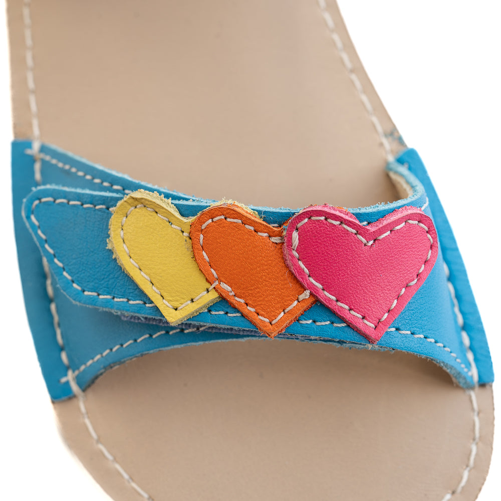A photo of the Zeazoo Ariel kids leather sandals. The sandals are blue in color and have velcro straps that go over the toes and around the ankles. The toe straps have a yellow, orange, and pink heart on them. The heart detail on the toe strap of the right shoe is shown close up on a white background. #color_light-blue-hearts