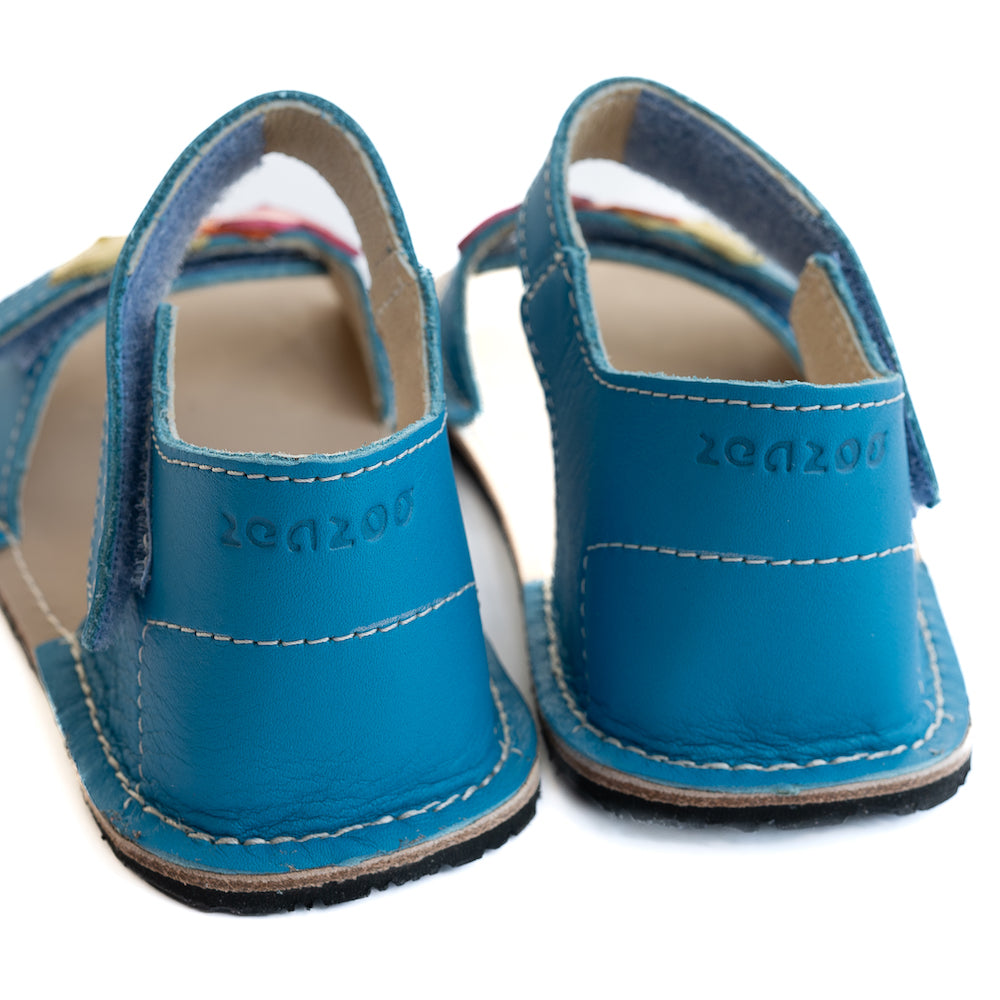 A photo of the Zeazoo Ariel kids leather sandals. The sandals are blue in color and have velcro straps that go over the toes and around the ankles. The toe straps have a yellow, orange, and pink heart on them. The sandals are shown together from behind on a white background, displaying the Zeazoo logo on the heels. #color_light-blue-hearts