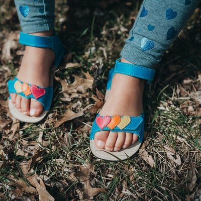 A photo of the Zeazoo Ariel kids leather sandals. The sandals are blue in color and have velcro straps that go over the toes and around the ankles. The toe straps have a yellow, orange, and pink heart on them. The sandals are shown together from above on a little girl wearing blue leggings with metallic hearts sitting on leafy grass. #color_light-blue-hearts