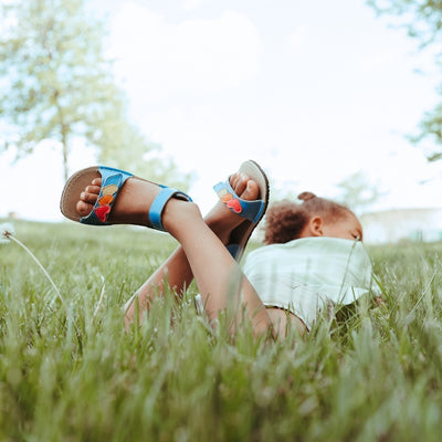 A photo of the Zeazoo Ariel kids leather sandals. The sandals are blue in color and have velcro straps that go over the toes and around the ankles. The toe straps have a yellow, orange, and pink hearts on them. Both shoes are shown on a dark-skinned little girl laying on her stomach in a grassy field with her feet crossed in the air.  #color_light-blue-hearts
