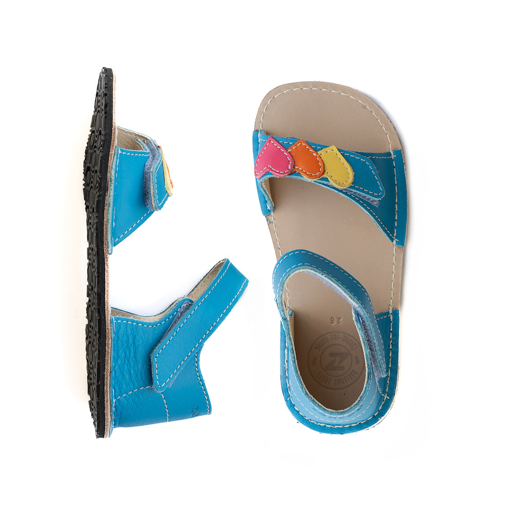 A photo of the Zeazoo Ariel kids leather sandals. The sandals are blue in color and have velcro straps that go over the toes and around the ankles. The toe straps have a yellow, orange, and pink heart on them. The left sandal is shown from the left side and the right sandal is shown from above on a white background. #color_light-blue-hearts#color_light-blue-hearts
