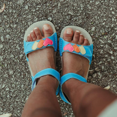A photo of the Zeazoo Ariel kids leather sandals. The sandals are blue in color and have velcro straps that go over the toes and around the ankles. The toe straps have a yellow, orange, and pink hearts on them. Both shoes are shown from above on a dark-skinned little girl standing on pavement. #color_light-blue-hearts