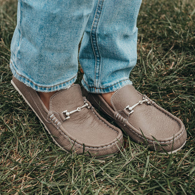 A photo of the Zeazoo Cheetah loafers made from a natural nappa leather upper on a tan Vibram sole. The loafers are stone in color and have a silver metal detail across the top of the foot. This photo shows both feet on a person standing in the grass. Slightly angled to the side. #color_stone