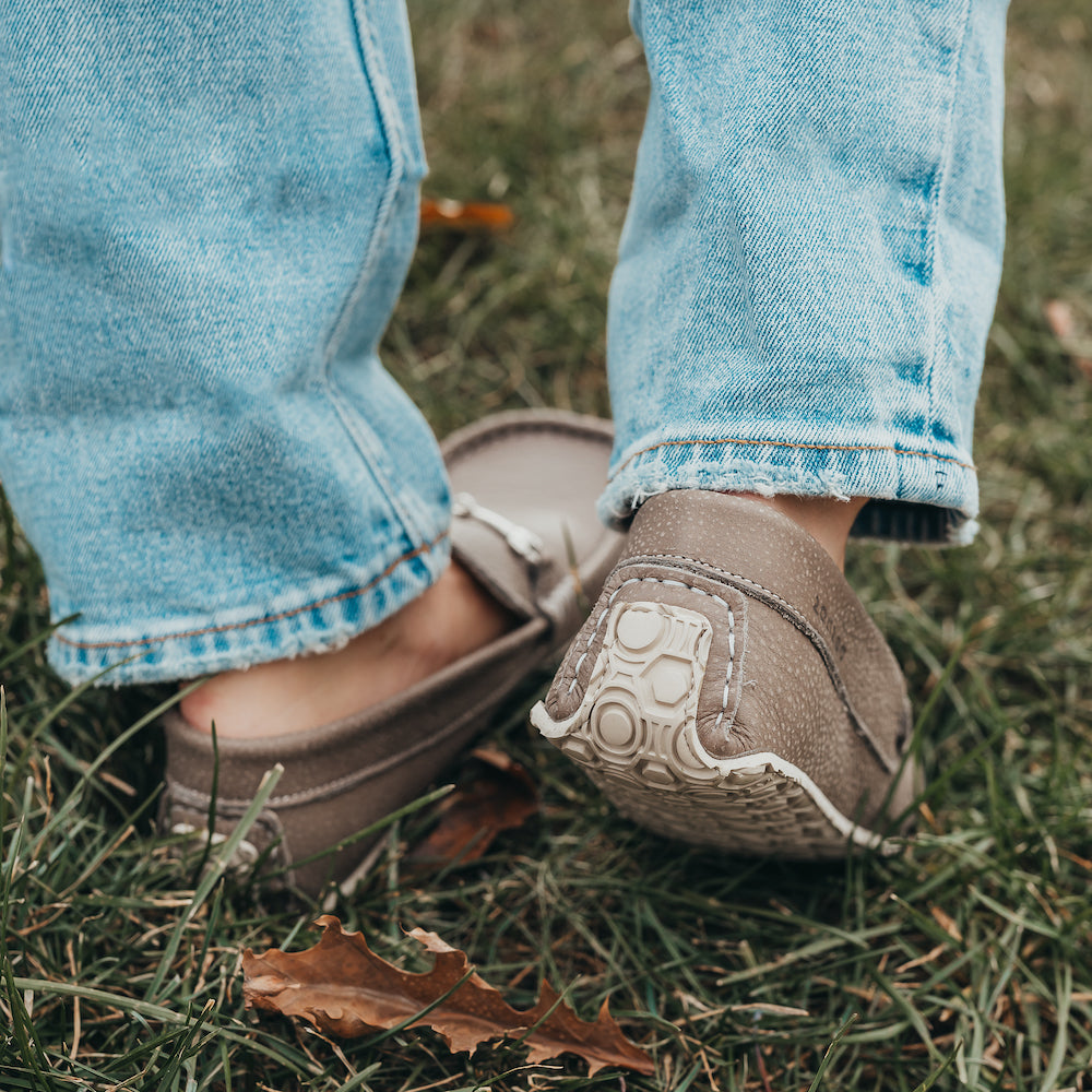 A photo of the Zeazoo Cheetah loafers made from a natural nappa leather upper on a tan Vibram sole. The loafers are stone in color and have a silver metal detail across the top of the foot. This photo shows both feet on a person standing in the grass. The heel detail of the right shoe is the focus of this image.  #color_stone