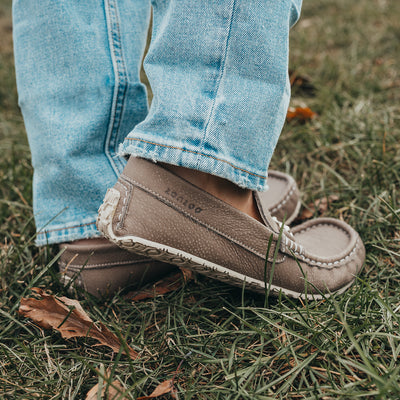 A photo of the Zeazoo Cheetah loafers made from a natural nappa leather upper on a tan Vibram sole. The loafers are stone in color and have a silver metal detail across the top of the foot. This photo shows both feet on a person standing in the grass. The side foot detail of the right shoe is the focus of this image. #color_stone