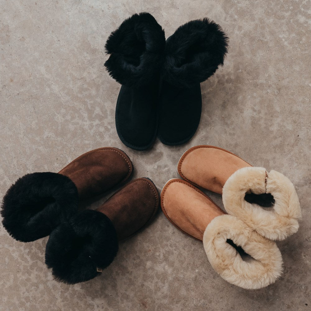 A photo of Zeezoo Dingo boots made with suede, sheepskin, and rubber soles. The boots are brown, black, and light brown in color with an ugg style look. Three pairs of boots are shown from the top down placed in a circle with the lining folded out and exposed against a cement floor. #color_black