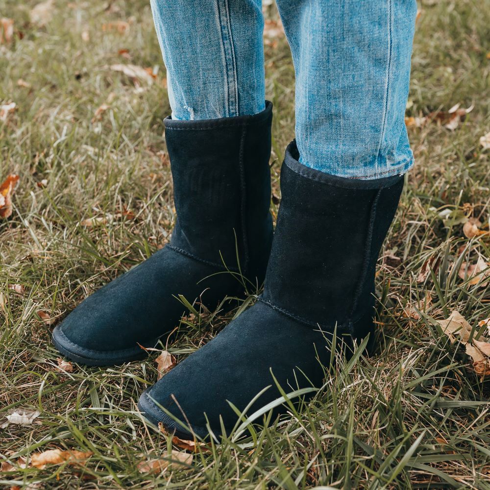 A photo of Zeezoo Dingo made with suede, sheepskin, and rubber soles. The boots are black in color with an ugg style look. Both boots are shown diagonally from the front left side on a woman’s feet, with a view of the woman’s shins down. The woman is wearing cropped blue jeans tucked into the boots, and is standing in grass. #color_black