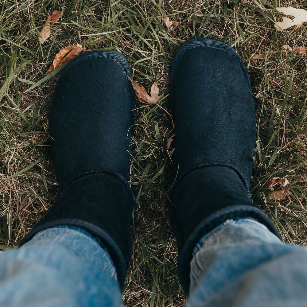 A photo of Zeezoo Dingo made with suede, sheepskin, and rubber soles. The boots are black in color with an ugg style look. Both boots are shown from above on a woman’s feet, with a view of the woman’s shins down. The woman is wearing cropped blue jeans tucked into the boots and is standing in grass. #color_black