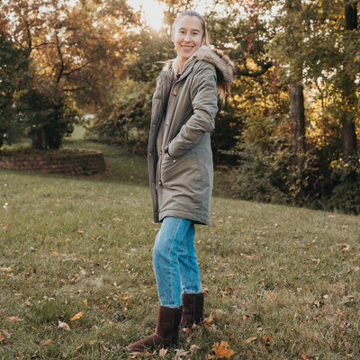 A photo of Zeezoo Dingo made with suede, sheepskin, and rubber soles. The boots are brown in color with tan soles and an ugg style look. Both boots are shown from the left side on a woman’s feet. The woman is wearing an army green parka and blue jeans tucked into the boots and is standing in grass with her hands in her pockets. #color_brown