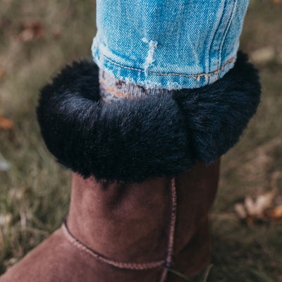 A photo of Zeezoo Dingo made with suede, sheepskin, and rubber soles. The boots are brown in color with tan soles and an ugg style look. The right boot is shown from the left side on a woman’s feet, with a view of her shins down and a close up of the cuff of the boot. The cuff of the boot is folded down to show the black sheepskin interior. The woman is wearing cropped blue jeans and the boots and is standing in grass. #color_brown