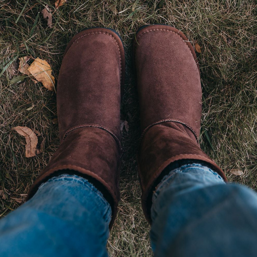 A photo of Zeezoo Dingo made with suede, sheepskin, and rubber soles. The boots are brown in color with an ugg style look. Both boots are shown from above on a woman’s foot with a view of her knees down. The woman is wearing blue jeans tucked into the boots and is standing in grass. #color_brown