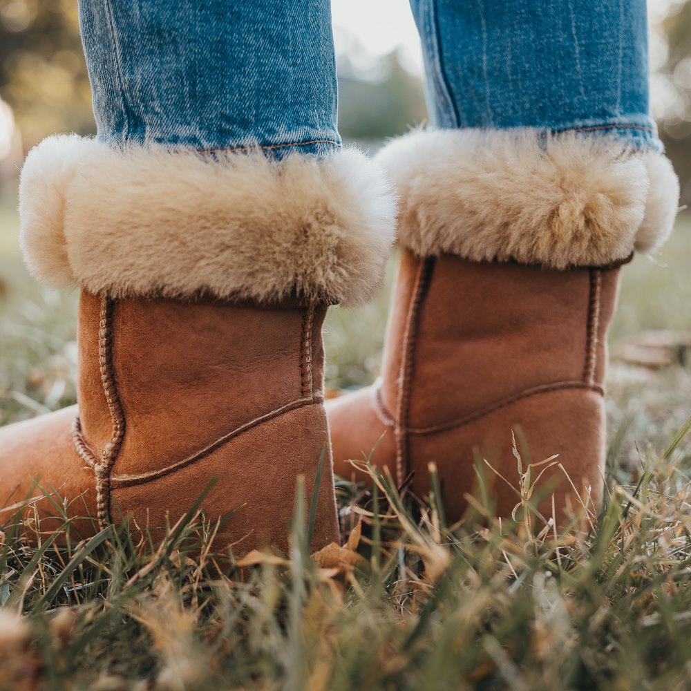 A photo of Zeezoo Dingo made with suede, sheepskin, and rubber soles. The boots are brown in color with tan soles and an ugg style look. Both boots are shown diagonally from the back left on a woman’s feet, with a view of her shins down. The cuff of the boots is folded down to show the sheepskin interior. The woman is wearing blue jeans tucked into the boots and is standing in grass. #color_chestnut
